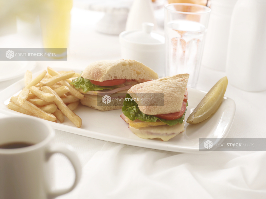 Peameal and melted cheese sandwich on a ciabatta bun with a pickle spear and french fries, white table setting