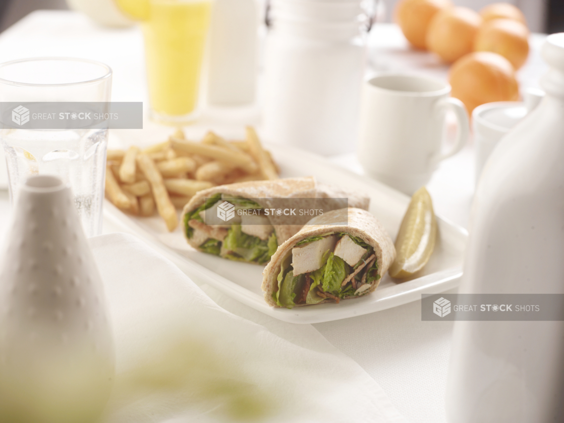 Salad wrap sandwich with chicken in two pieces on a plate with a pickle and french fries, white table setting