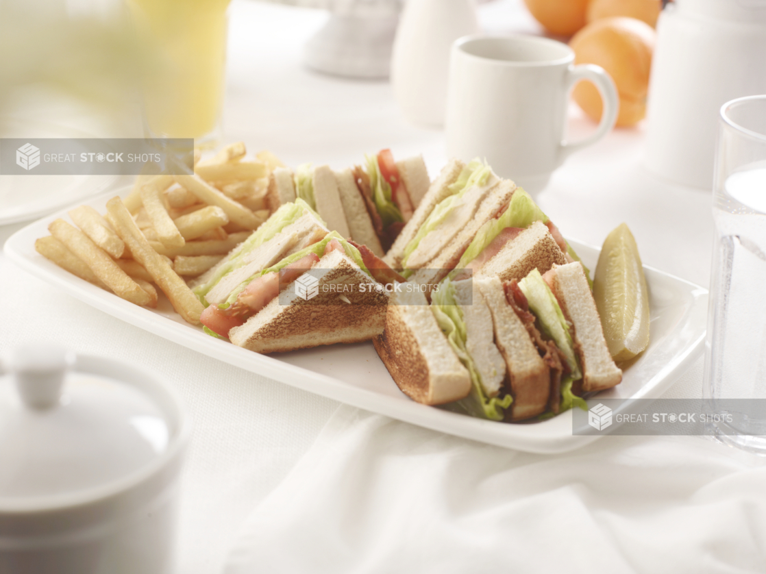 Chicken club sandwich, quartered, with french fries, on a square white plate in an all-white table setting