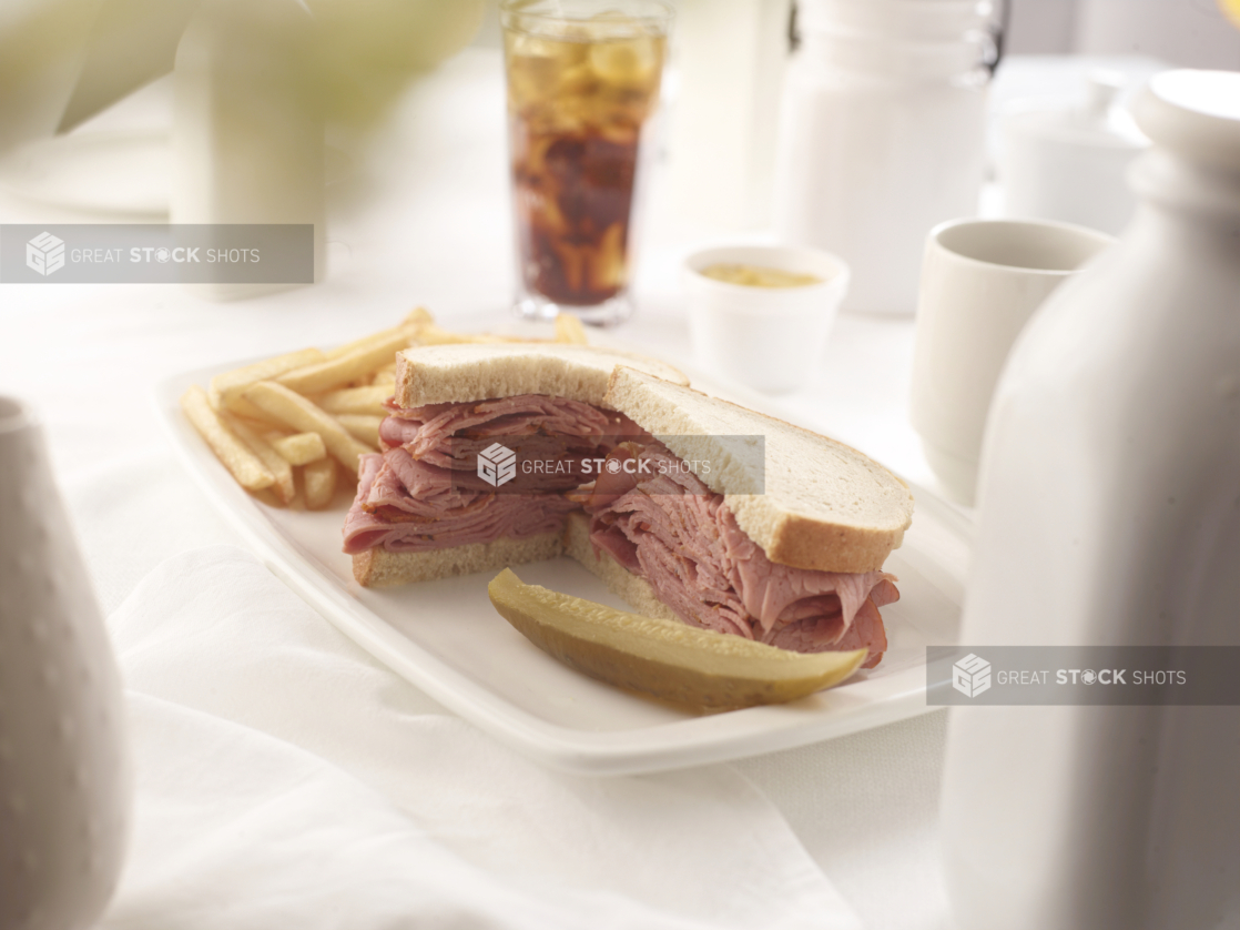 Pastrami sandwich on rye bread with a pickle and french fries, white table setting