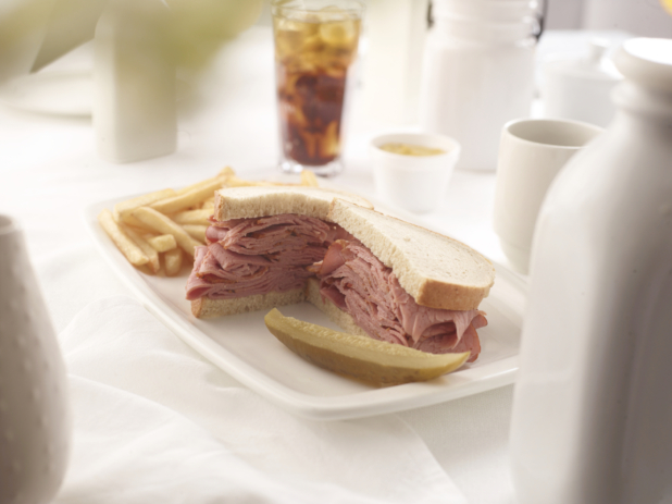 Pastrami sandwich on rye bread with a pickle and french fries, white table setting