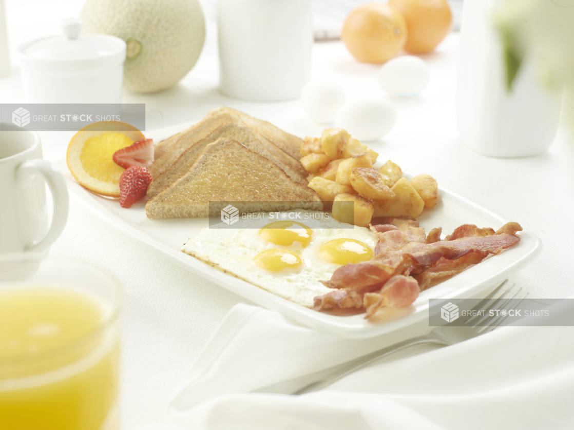 Breakfast of 3 eggs sunny side up with bacon, brown toast, homefries, and fruit, white table setting