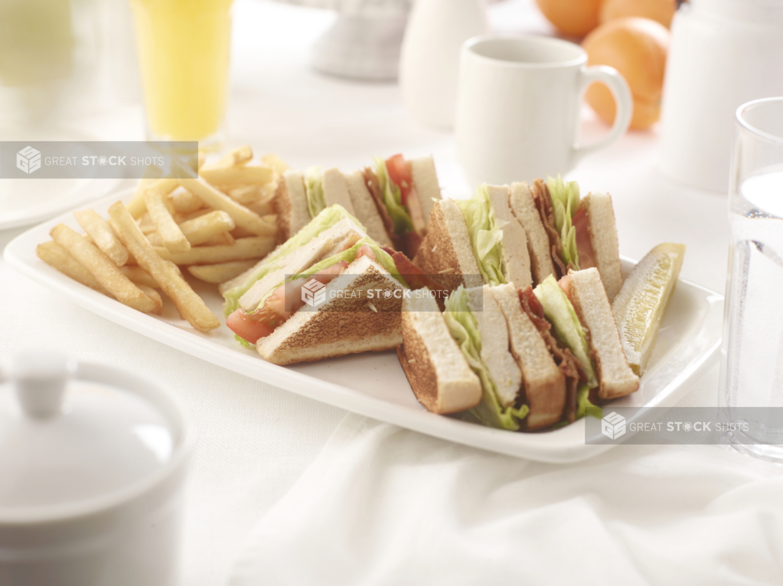 Chicken club sandwich, quartered, with french fries, on a square white plate in an all-white table setting