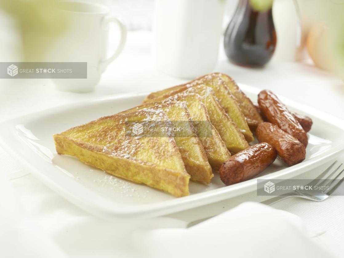 French toast with breakfast sausages on a square white plate in an all-white table setting