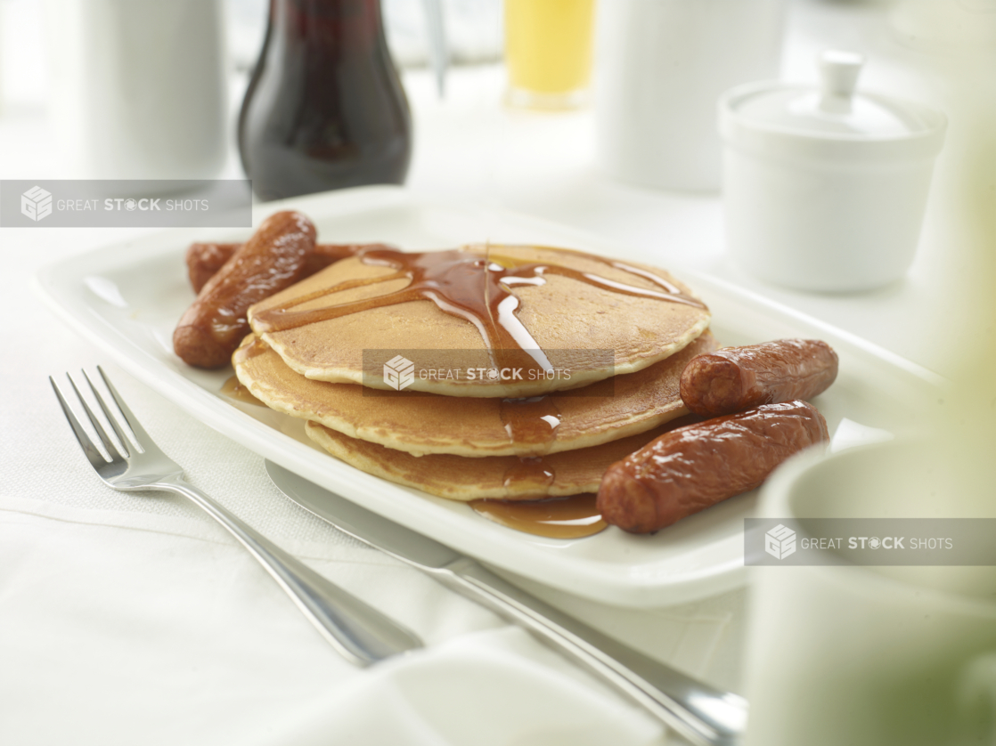 A Platter with a Stack of Buttermilk Pancakes with Maple Syrup and Breakfast Sausages on a White Table Cloth Surface in a Restaurant Setting