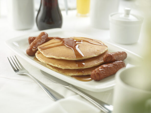 A Platter with a Stack of Buttermilk Pancakes with Maple Syrup and Breakfast Sausages on a White Table Cloth Surface in a Restaurant Setting