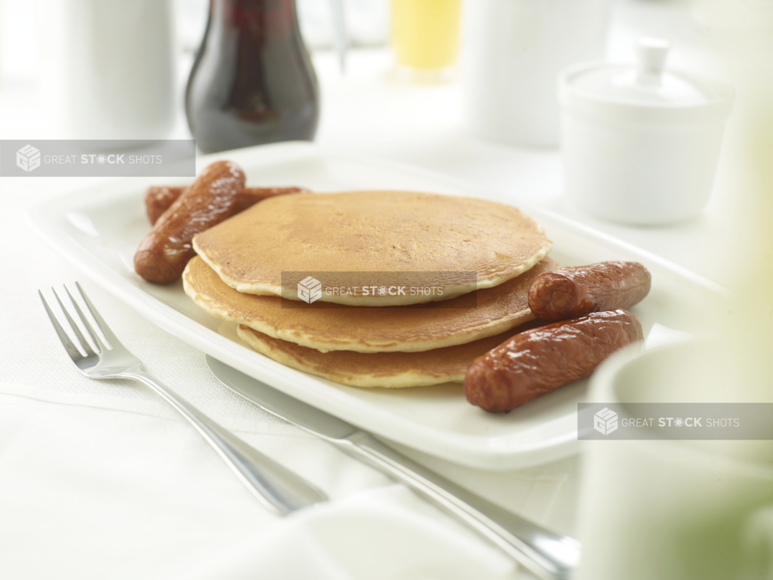 A Platter with a Stack of Plain Buttermilk Pancakes and Breakfast Sausages on a White Table Cloth Surface in a Restaurant Setting