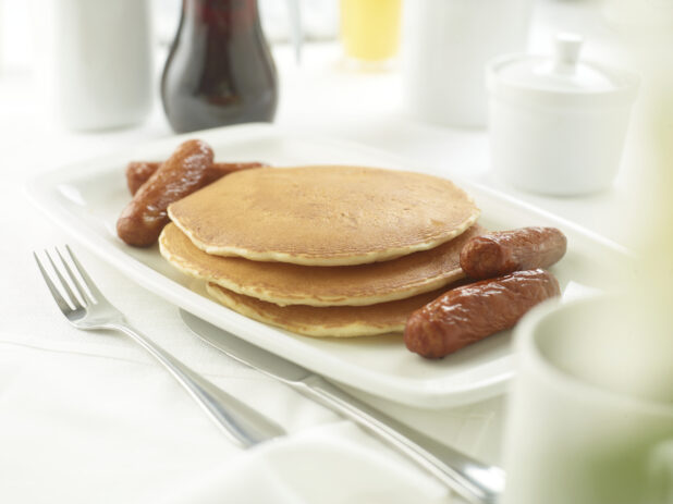 A Platter with a Stack of Plain Buttermilk Pancakes and Breakfast Sausages on a White Table Cloth Surface in a Restaurant Setting