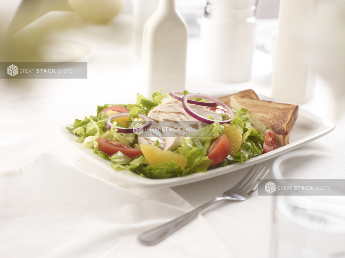 California-style romaine salad with grilled chicken, red onions, oranges, and tomato wedges with white toast on the side, white table setting