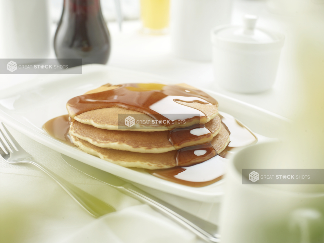 A Stack of Buttermilk Pancakes with Maple Syrup in a White Platter on a White Table Cloth Surface in a Restaurant Setting