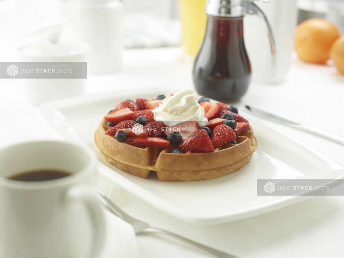 Belgian waffle with fresh strawberries and blueberries topped with whipped cream in an all-white table setting