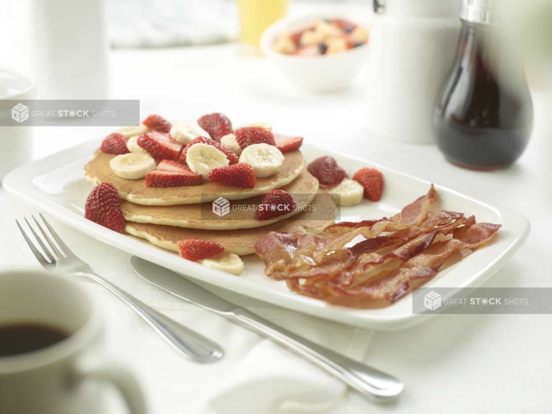 A Platter of Strawberries and Banana Pancakes with a Side of Bacon on a White Table Cloth in a Restaurant Setting