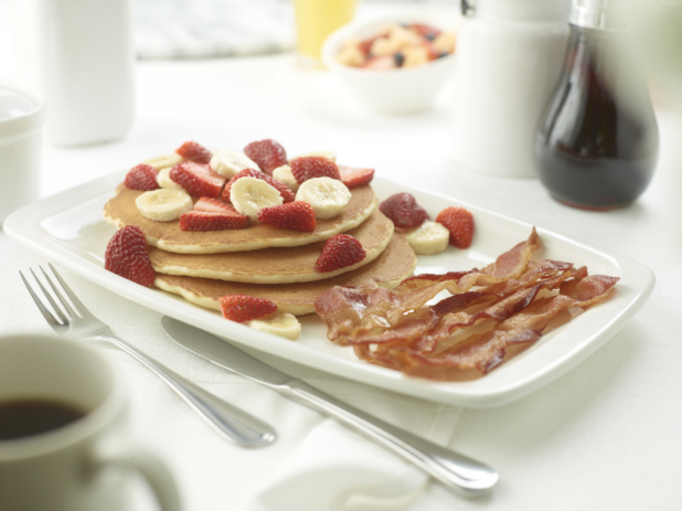 A Platter of Strawberries and Banana Pancakes with a Side of Bacon on a White Table Cloth in a Restaurant Setting