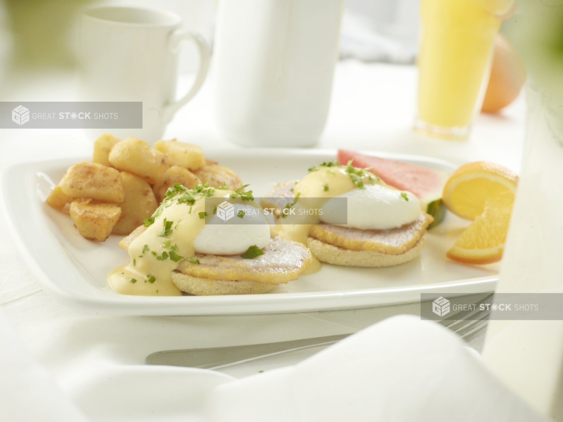 Classic Eggs Benedict with homefries and fresh fruit in an all-white setting, white background