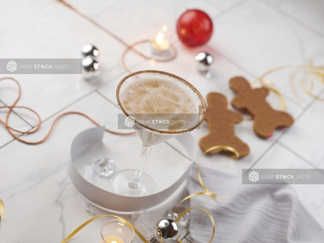 Eggnog martini on a white tabletop with gingerbread men and silver, red, and gold Christmas decorations