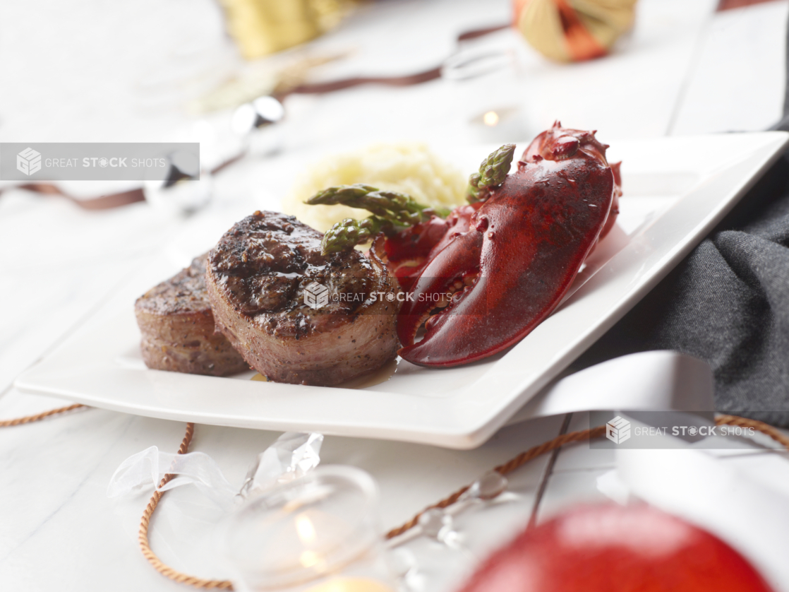 Close Up of a Square White Plate of Steak Medallions, Asparagus, Mashed Potatoes and Lobster Claws in an Indoor Setting with Holiday Decorations