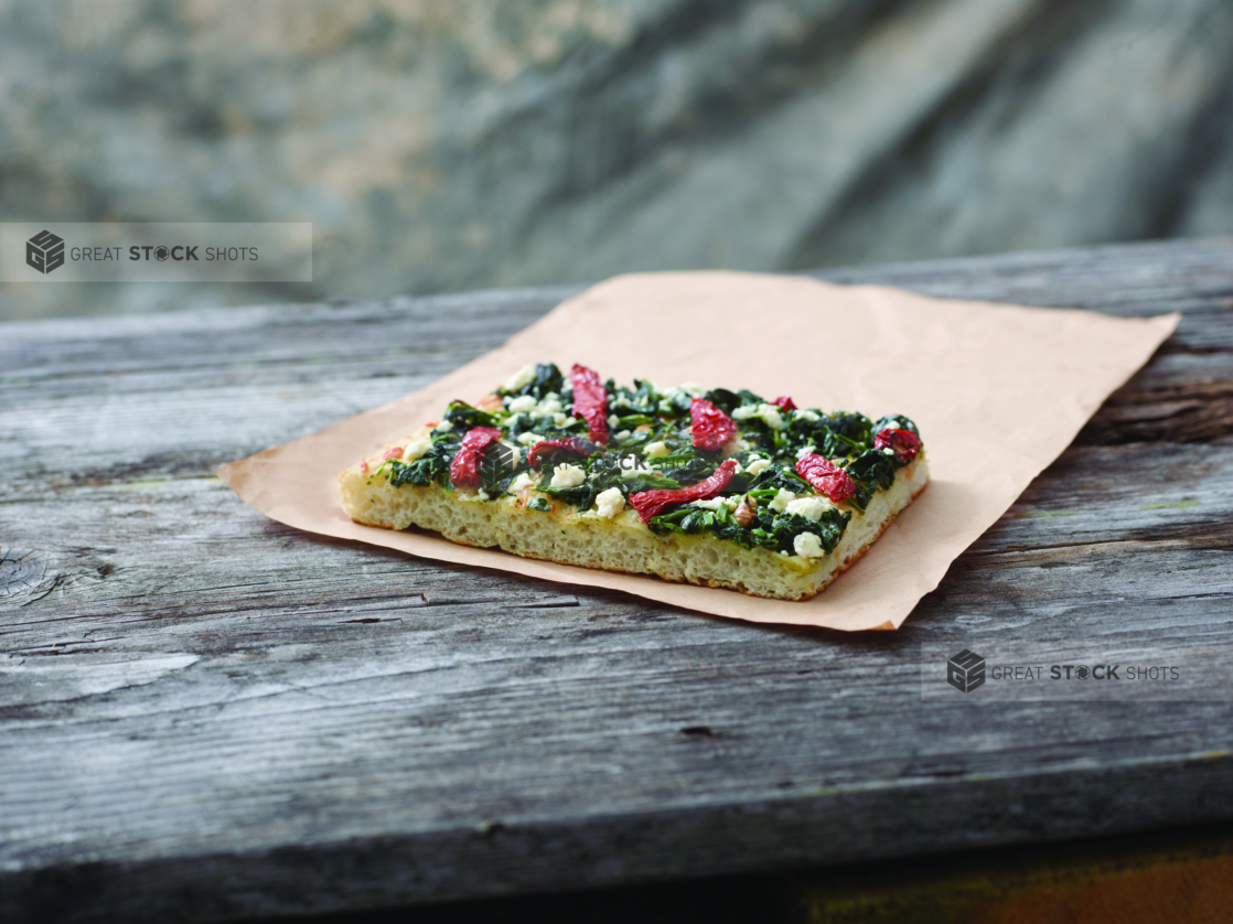 A Romano Pizza al Taglio Slice with Sautéed Spinach, Sun-dried Tomatoes and Feta Cheese on Kraft Paper on a Weathered Wooden Table