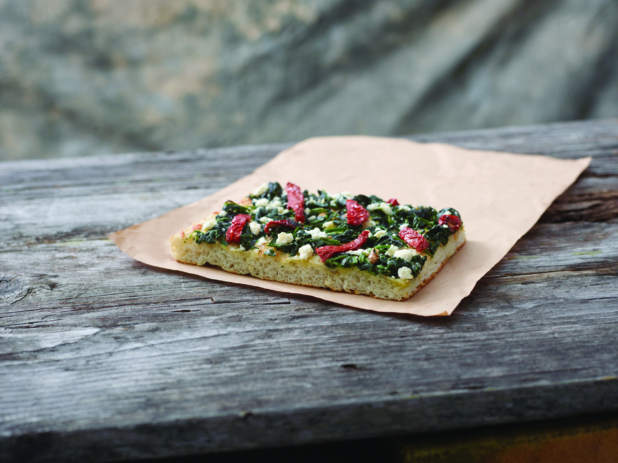 A Romano Pizza al Taglio Slice with Sautéed Spinach, Sun-dried Tomatoes and Feta Cheese on Kraft Paper on a Weathered Wooden Table