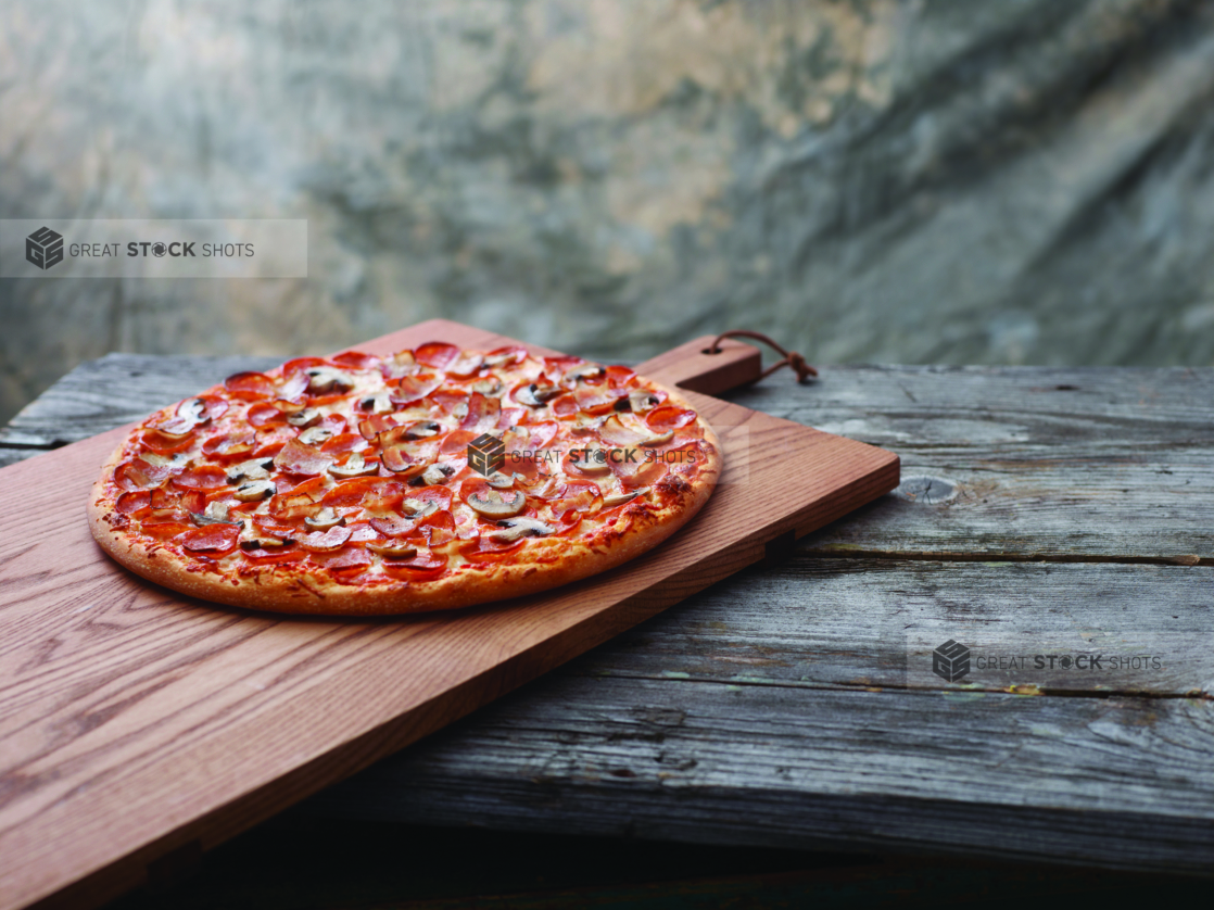 A Canadian Pizza with Pepperoni, Bacon and Mushrooms on a Wooden Cutting Board on a Aged Wooden Table Against a Brown Canvas Background