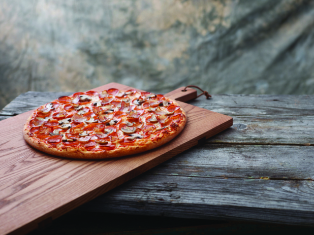 A Canadian Pizza with Pepperoni, Bacon and Mushrooms on a Wooden Cutting Board on a Aged Wooden Table Against a Brown Canvas Background