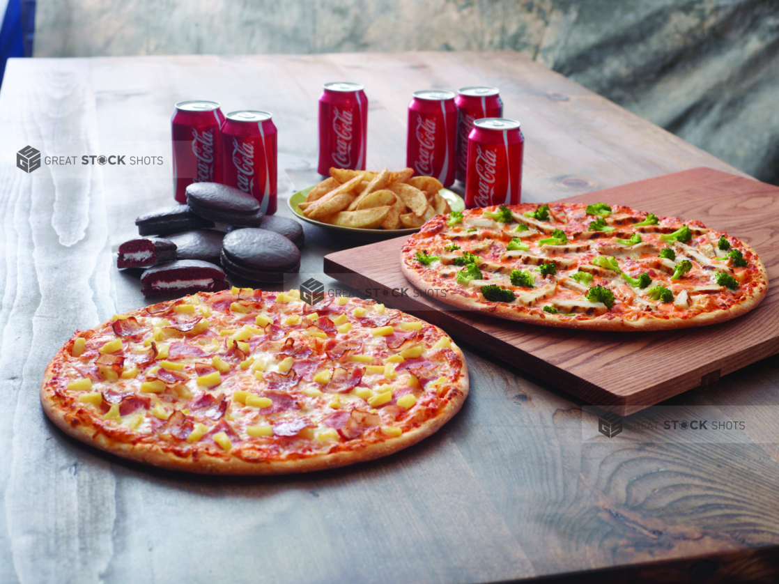 Family Pizza Special with a Hawaiian Pizza and Chicken Broccoli Pizza, Chocolate Cakes, Potato Wedges and a 6-Pack of Soda on a Dark Wooden Surface