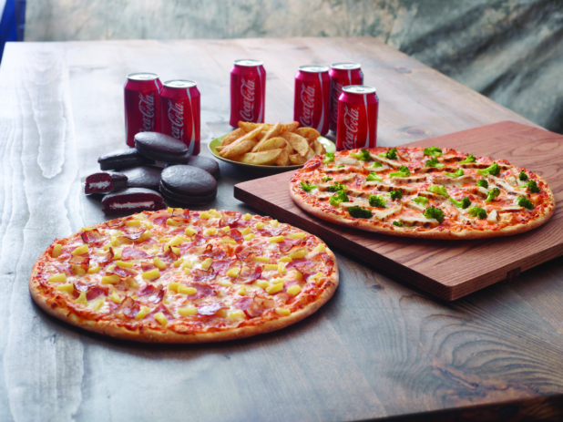 Family Pizza Special with a Hawaiian Pizza and Chicken Broccoli Pizza, Chocolate Cakes, Potato Wedges and a 6-Pack of Soda on a Dark Wooden Surface