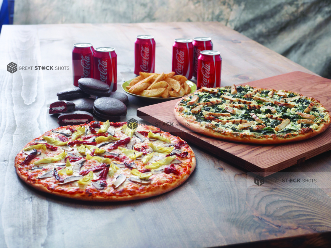 Family Pizza Special with a Chicken Florentine Pizza and a 3-Topping Vegetarian Pizza, Chocolate Cakes, Potato Wedges and a 6-Pack of Soda on a Dark Wooden Surface