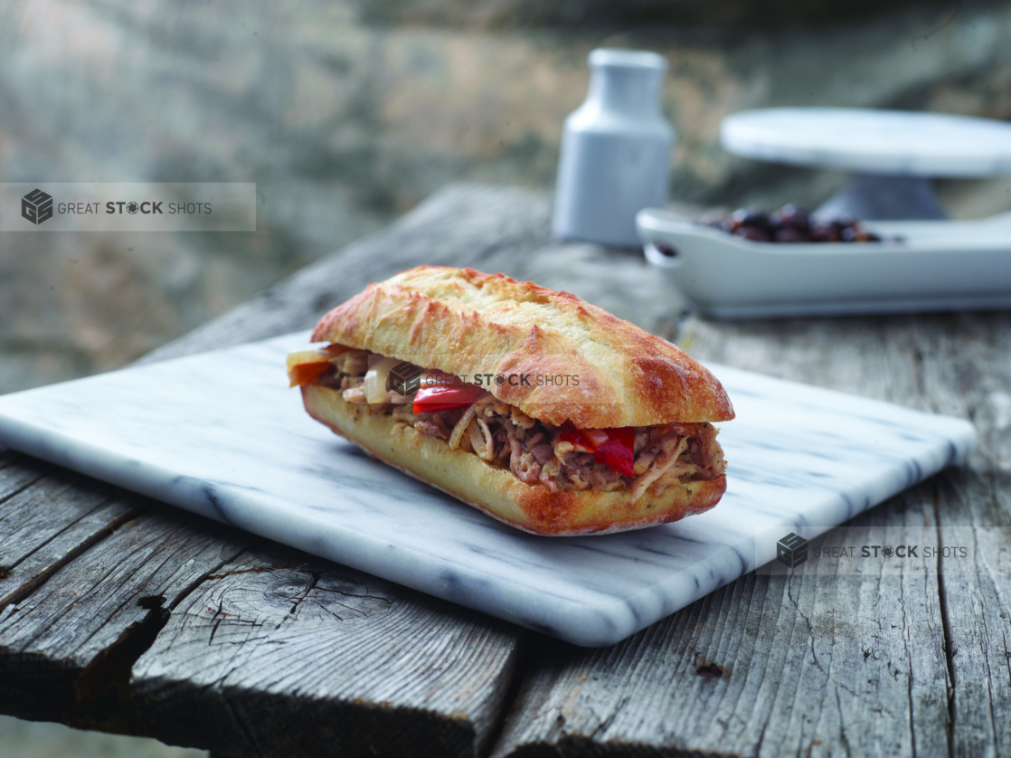 Italian Sandwich with Porchetta and Hot Banana Peppers in a Toasted Ciabatta Bun on a Marble Cutting Board on an Aged Wooden Table