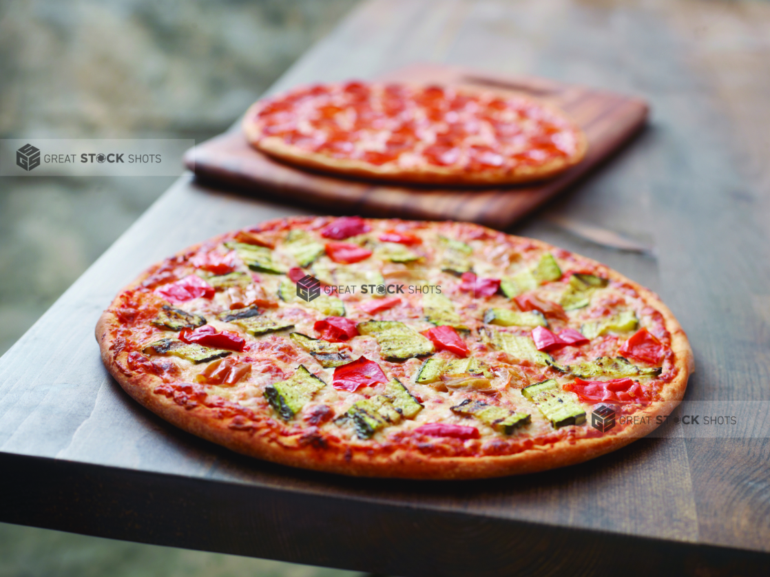 A Pizza with Grilled Zucchini and Hot Banana Peppers and a Pepperoni Pizza on a Wooden Cutting Board on a Dark Wood Table Against a Brown Canvas Background