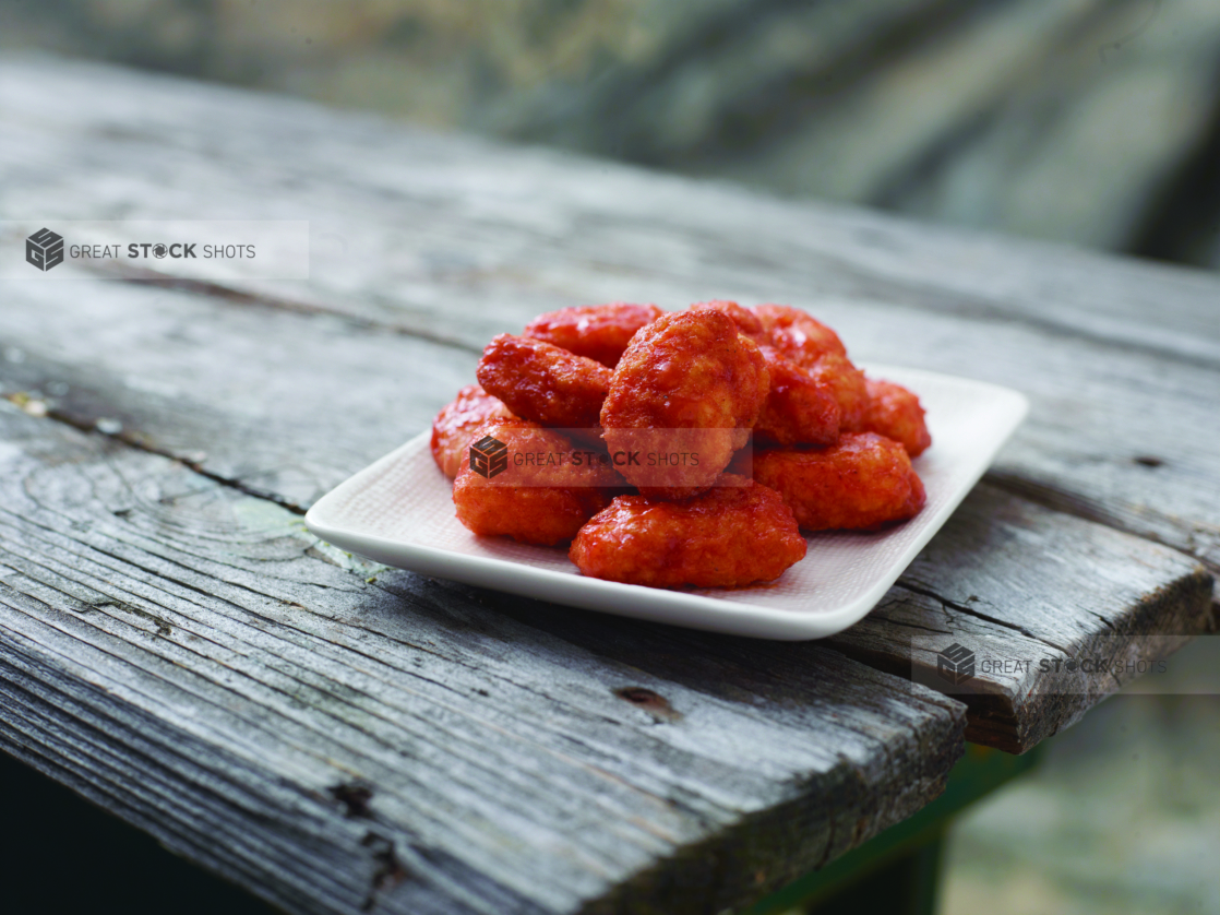 A Close Up of a Dish of Sauced Boneless Chicken Wings on a Weathered Wooden Table Against a Canvas Background