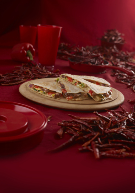 Steak quesadilla stacked in wedges on a round wood board in an all-red setting surrounded by dried red chilis