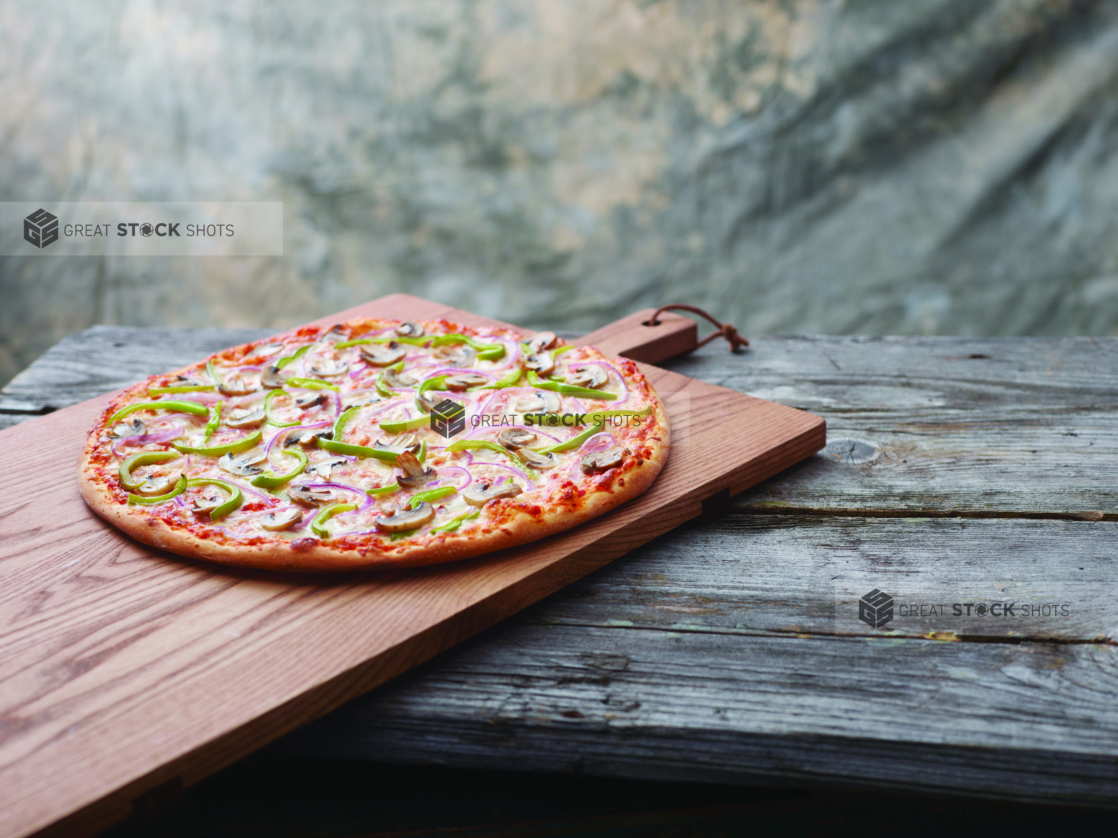 A Pizza with Green Onions, Mushrooms and Red Onions on a Wooden Cutting Board on a Aged Wooden Table Against a Brown Canvas Background