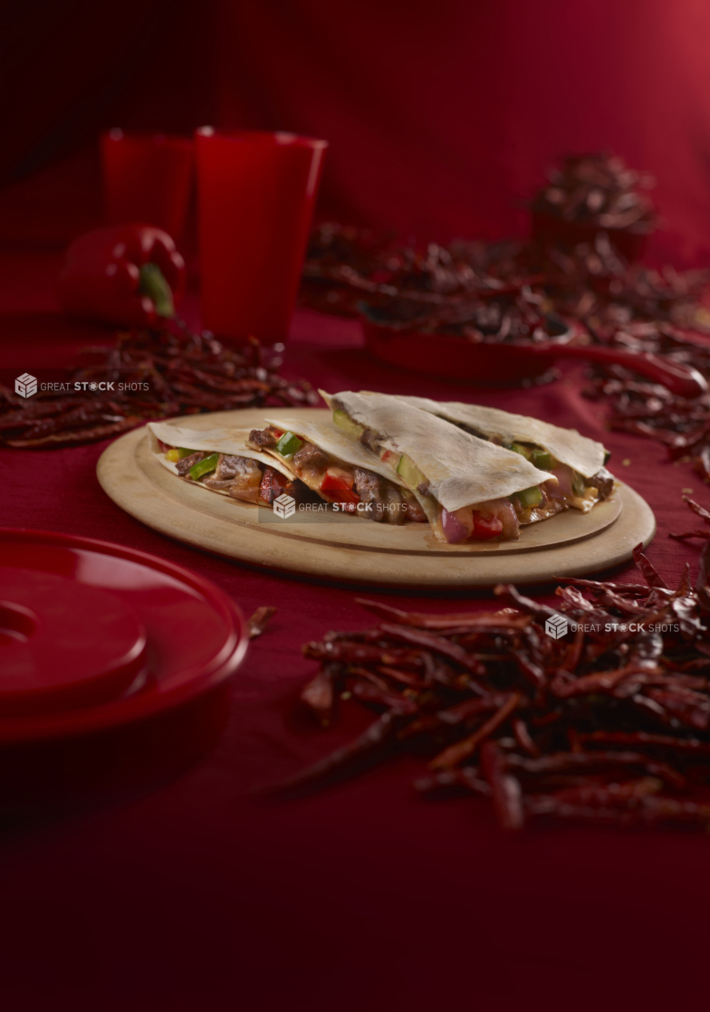 Steak quesadilla stacked in wedges on a round wood board in an all-red setting surrounded by dried red chilis