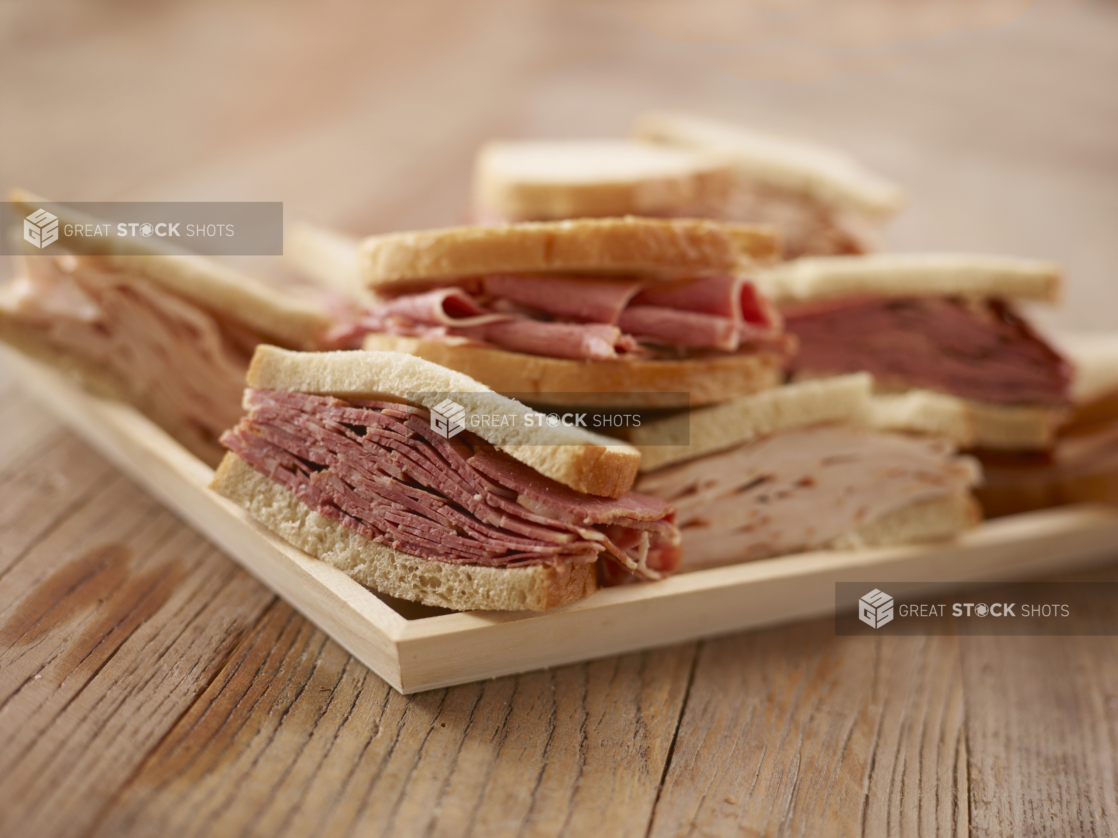 Close Up of a Wooden Catering Platter of Pastrami and Roast Turkey Sandwiches on White Rye Bread on a Weathered Wooden Surface