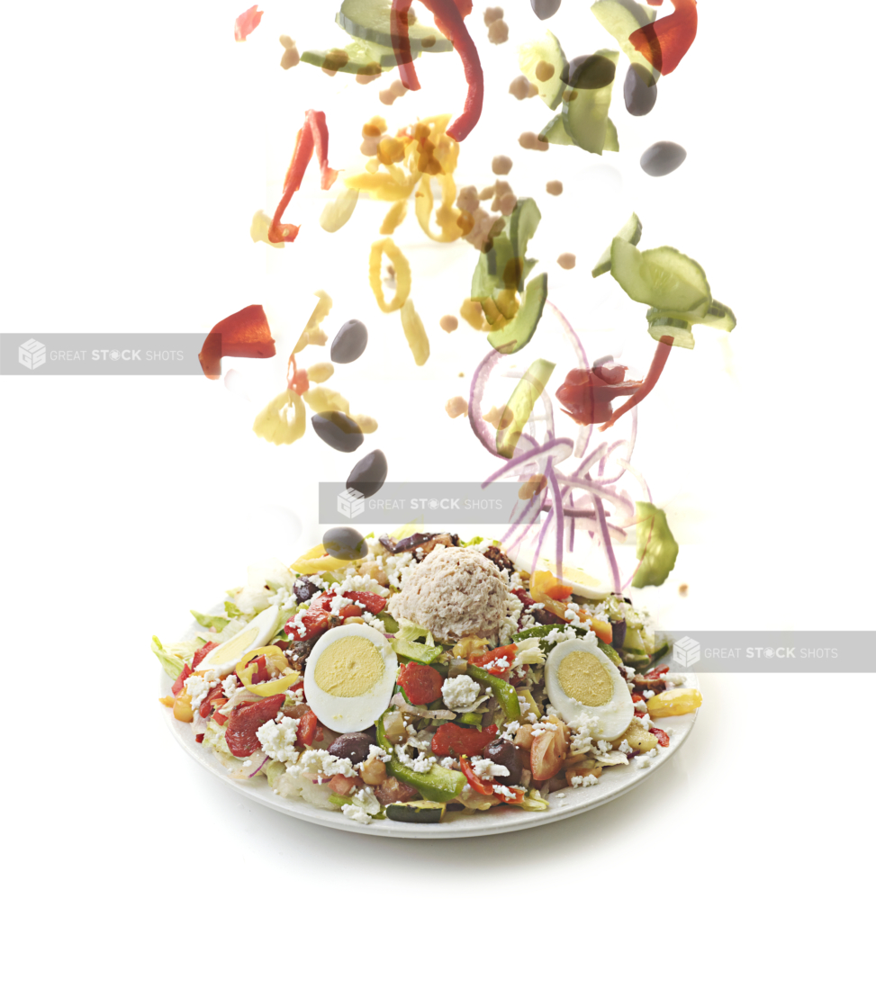 Fresh Ingredients Falling onto a Healthy Salad of Freshly Chopped Vegetables, Boiled Eggs, Feta Cheese and a Scoop of Tuna on a Round White Plate, Isolated on a White Background