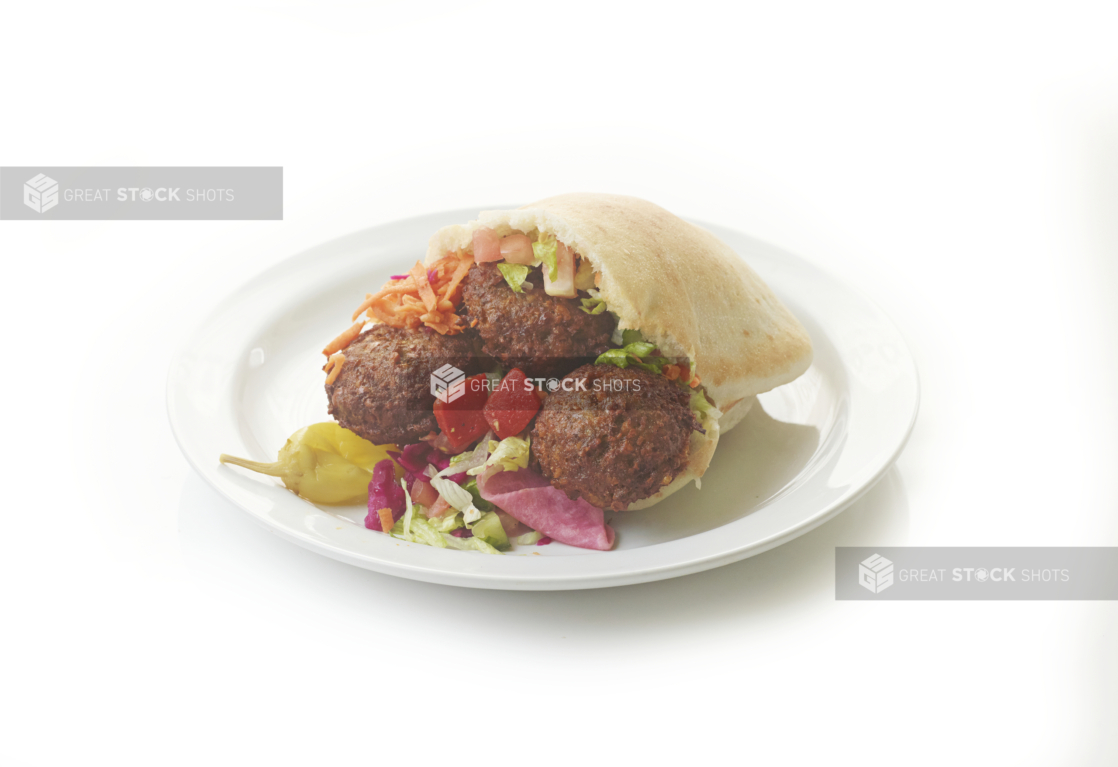 A White Pita Pocket Stuffed with Deep Fried Falafel Balls, Shredded Vegetables and Pickled Veggies with a Peppercini on a Round White Plate, Isolated on a White Background