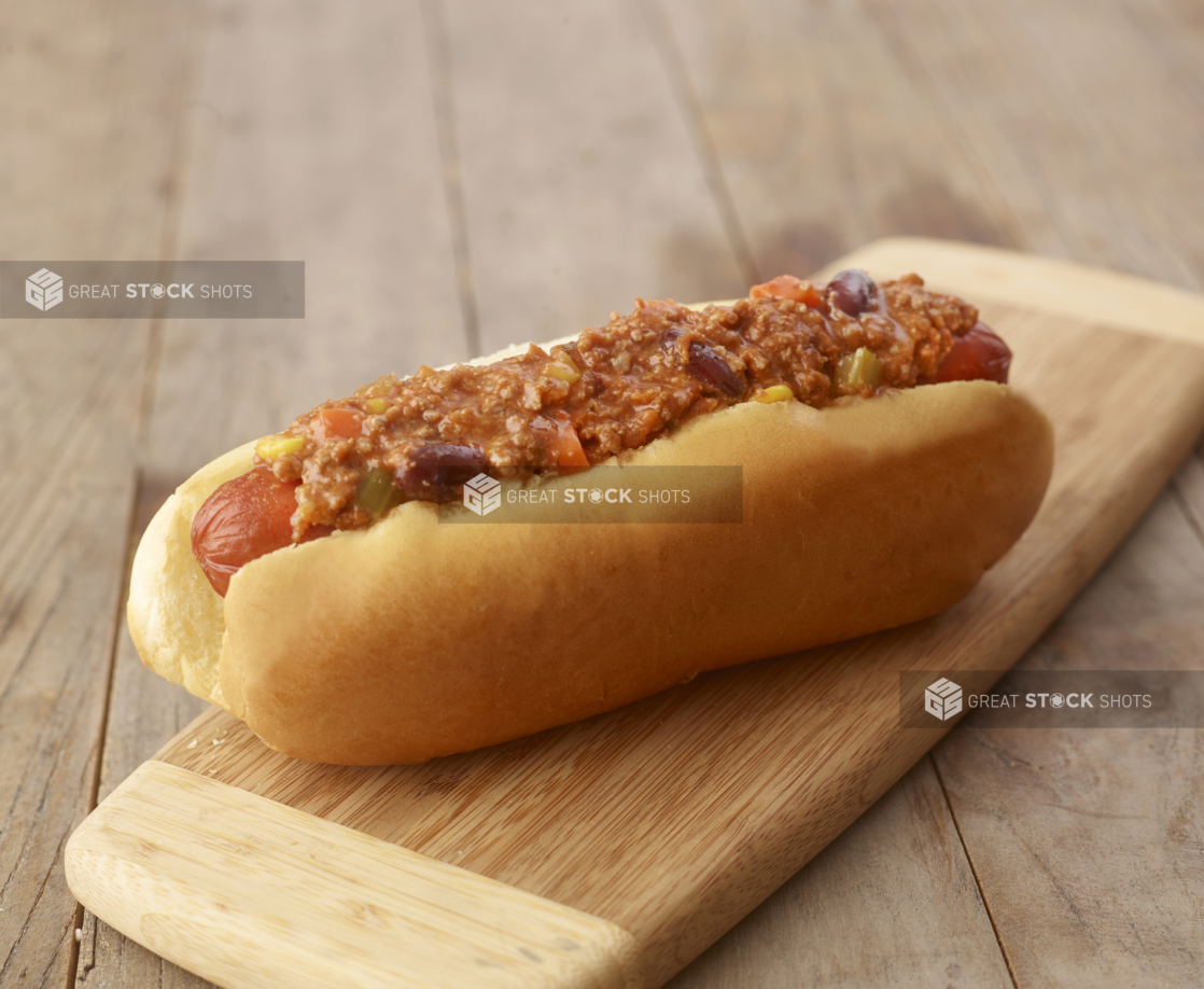 Close Up of a Chilli Hot Dog on a Small Wooden Cutting Board on a Rustic Wood Surface