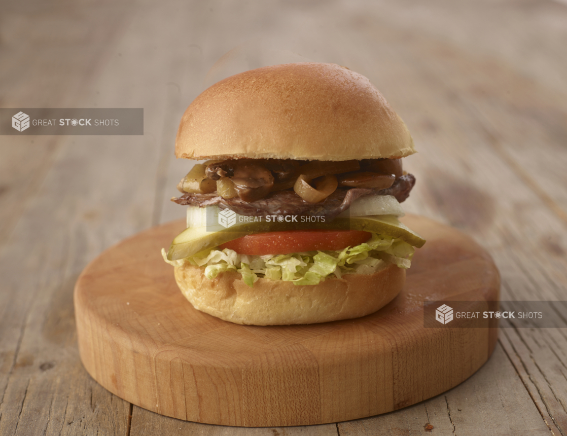 Close up of a Steak, Mushroom and Onion Burger with Fresh Vegetable Toppings on a Wooden Cutting Board on a Weathered Wood Surface