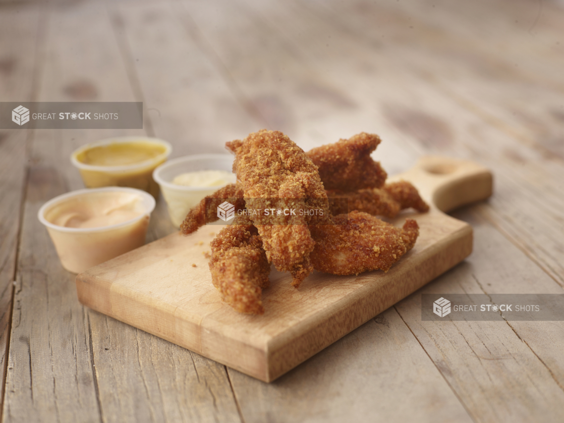 Deep Fried Breaded Chicken Tenders on a Wooden Cutting Board with a Trio of Dipping Sauces on a Weathered Wooden Surface