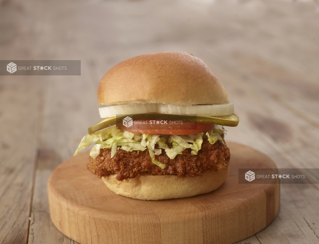 Close up of a Fried Chicken Sandwich with Fresh Vegetable Toppings on a Wooden Cutting Board on a Weathered Wood Surface