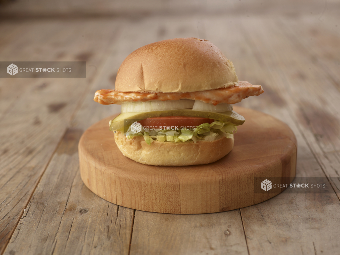 Close up of a Grilled Chicken Sandwich with Fresh Vegetable Toppings on a Wooden Cutting Board on a Weathered Wood Surface