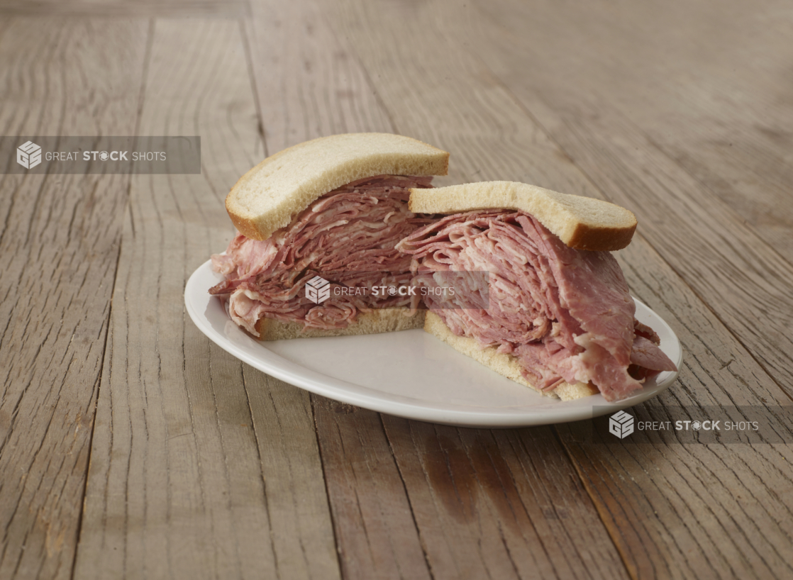 Halved pastrami sandwich on sliced rye bread on a white plate, wood tabletop