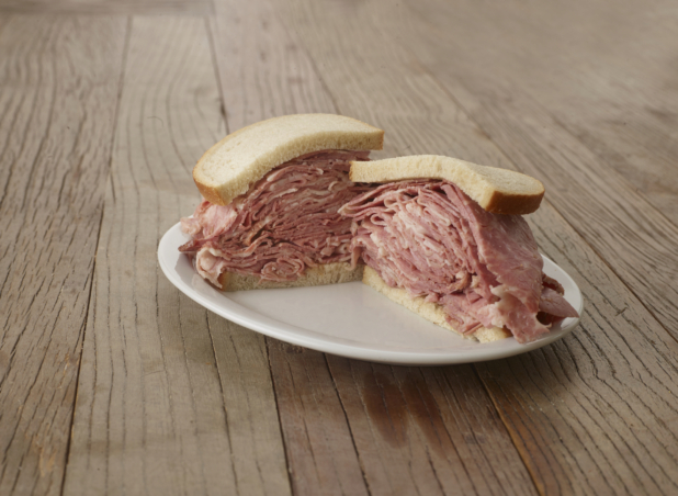 Halved pastrami sandwich on sliced rye bread on a white plate, wood tabletop