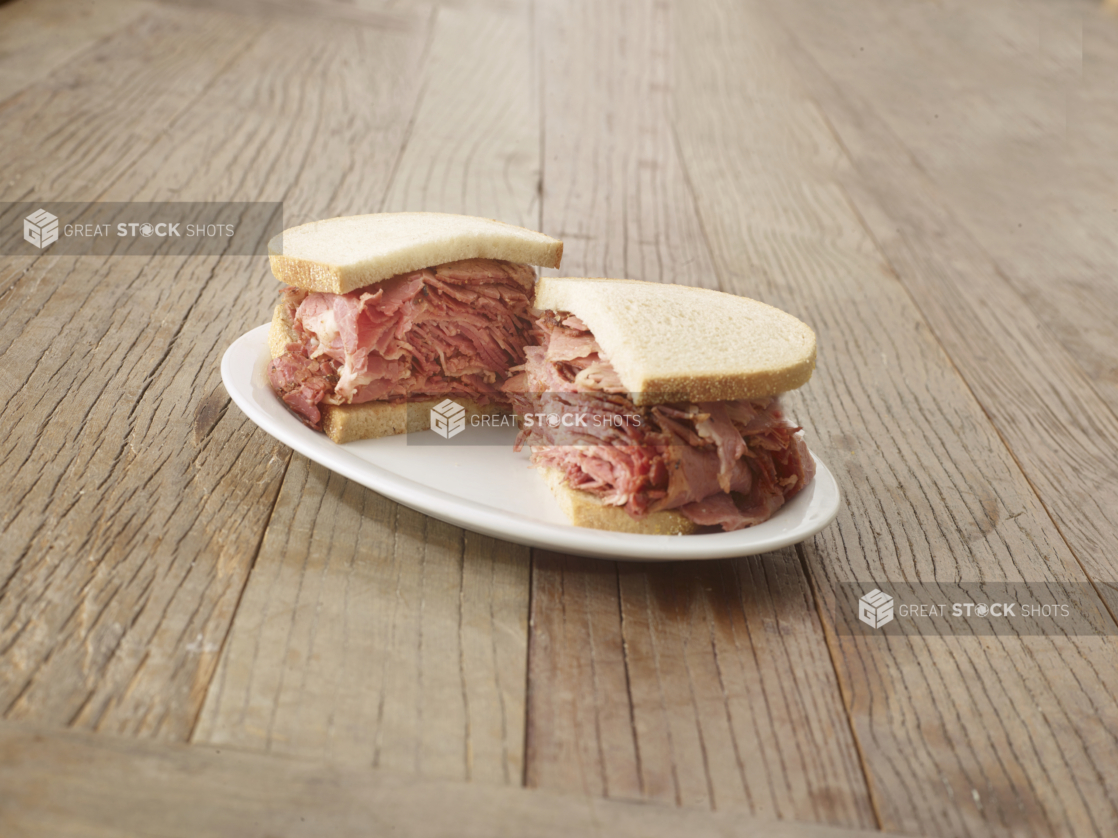 Halved pastrami sandwich on sliced rye bread on a white plate, wood tabletop