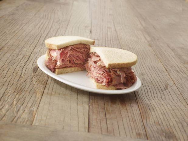 Halved pastrami sandwich on sliced rye bread on a white plate, wood tabletop