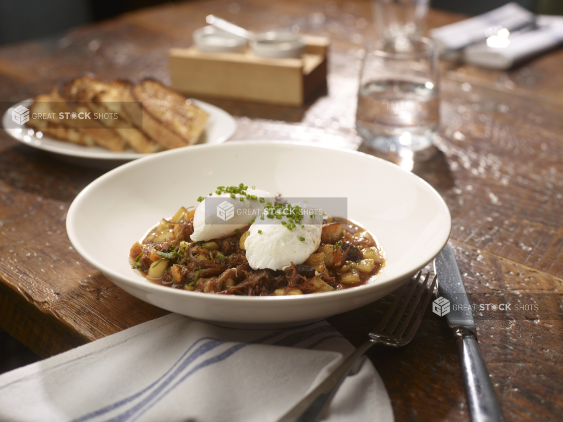 A White Ceramic Bowl of Beef Short Rib Hash Topped with Two Poached Eggs on a Wooden Table in a Restaurant Setting