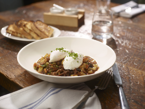 A White Ceramic Bowl of Beef Short Rib Hash Topped with Two Poached Eggs on a Wooden Table in a Restaurant Setting