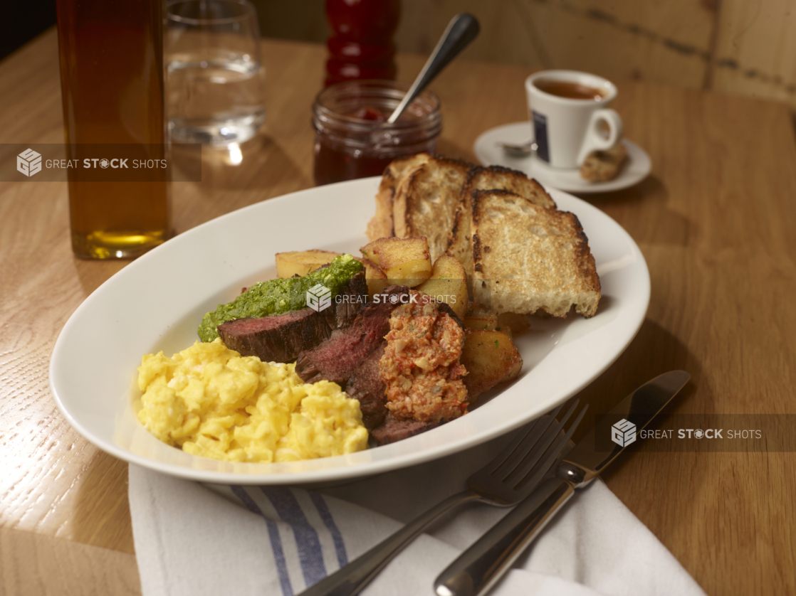 A breakfast plate of scrambled eggs, brisket, potatoes and toast with an espresso in background