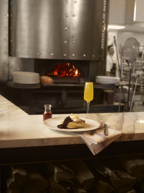 Plate of pancakes with blueberry compote and cream and a mimosa in a tall stemmed glass on a marble bar in front of a pizza oven