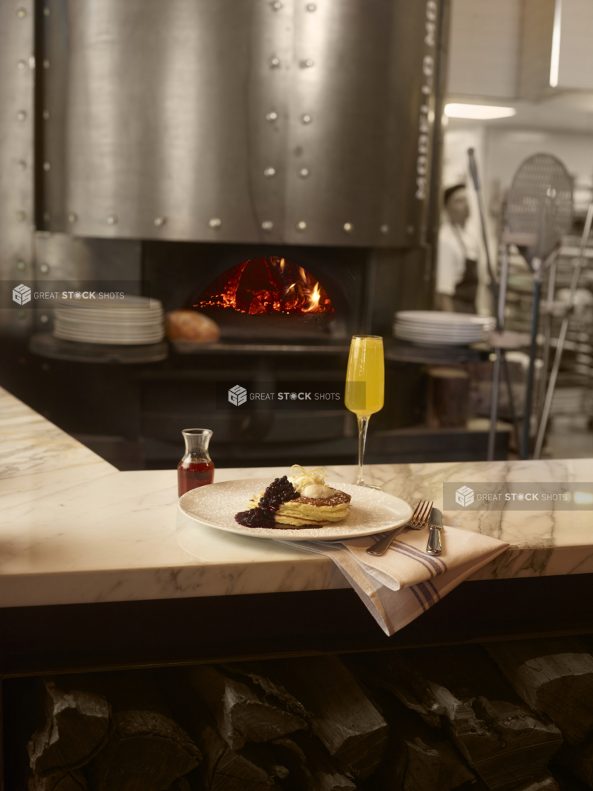 Plate of pancakes with blueberry compote and cream and a mimosa in a tall stemmed glass on a marble bar in front of a pizza oven